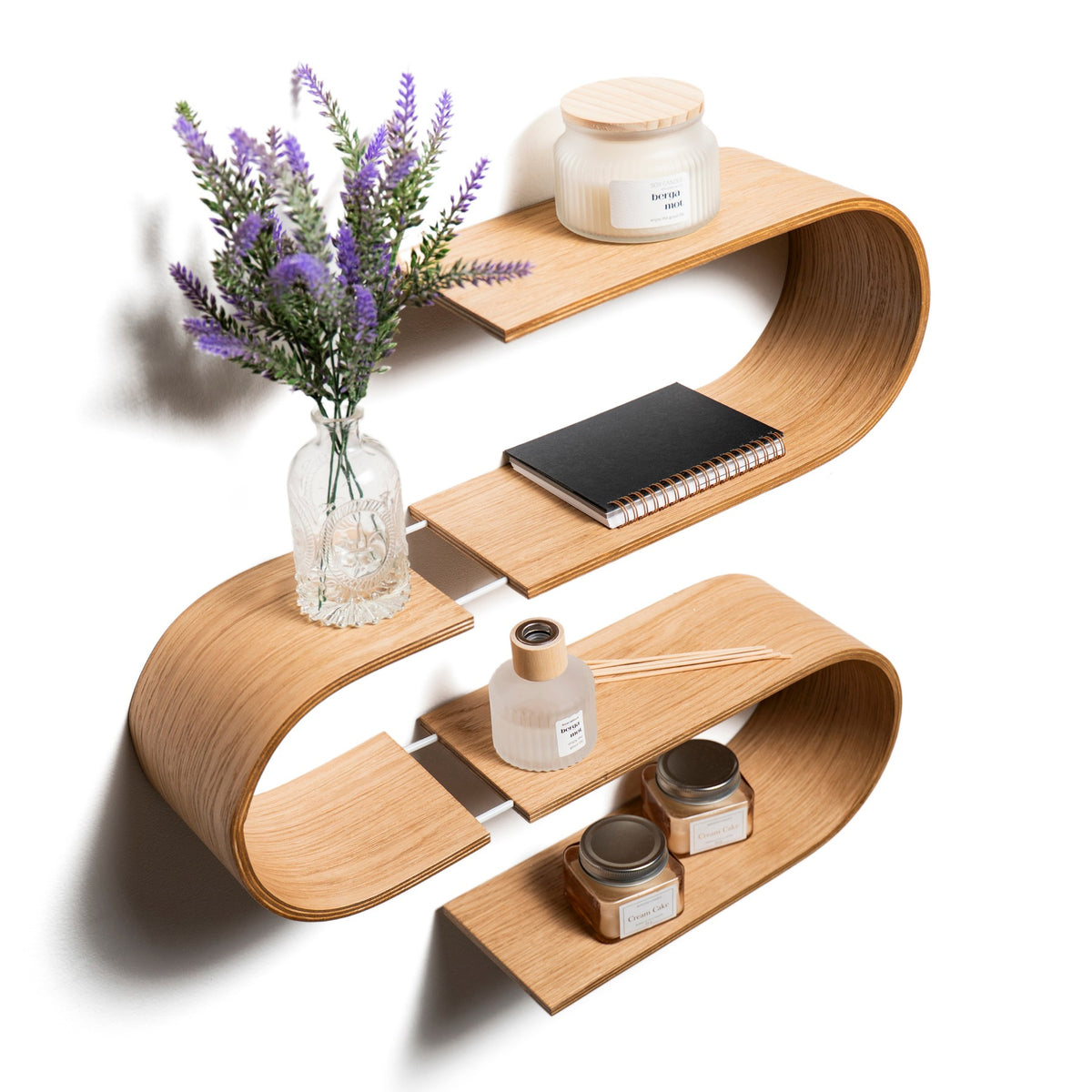 Angled view of the S-shaped wooden wall shelf styled with a vase, candle and notebook